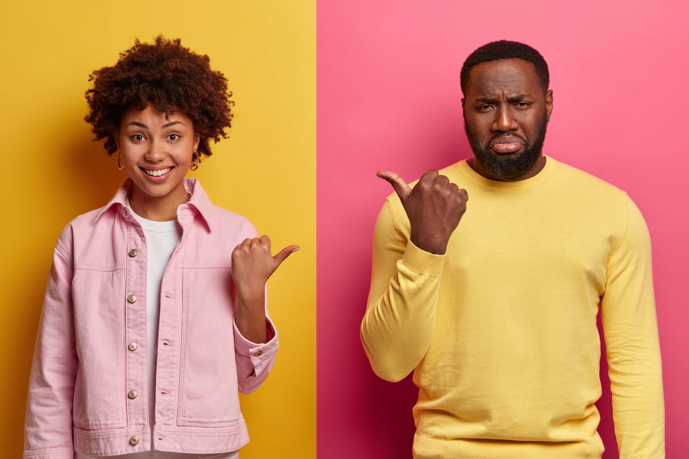 A woman and a man pointing their thumbs at each other, the woman is smiling, while the man is pouting.