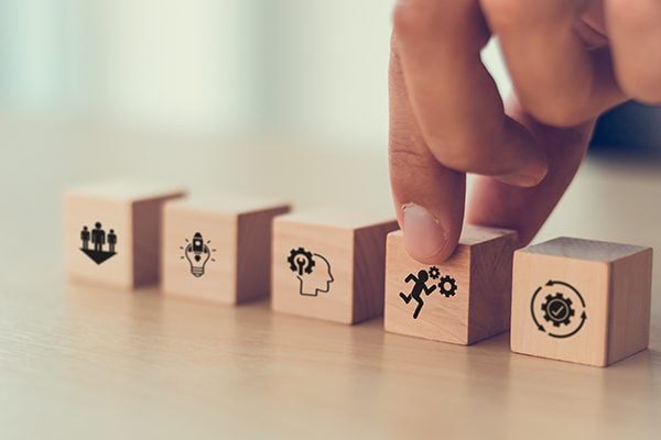 wooden blocks lined up evenly on a table with icons on each and a person touching a block with the icon on a running person with two gears