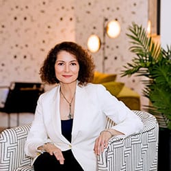 Kristina Dipalo ACC headshot photo, smiling woman with dark curly hair wearing a white blazer and black dress sitting