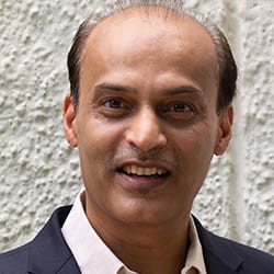 Sandeep Jain PCC headshot photo, smiling man in blazer and collared shirt in front of white wall