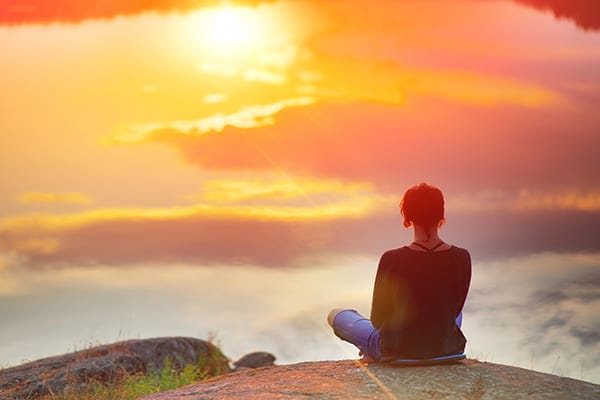 person sitting on the edge of a mountain at sunrise meditating for wellbeing